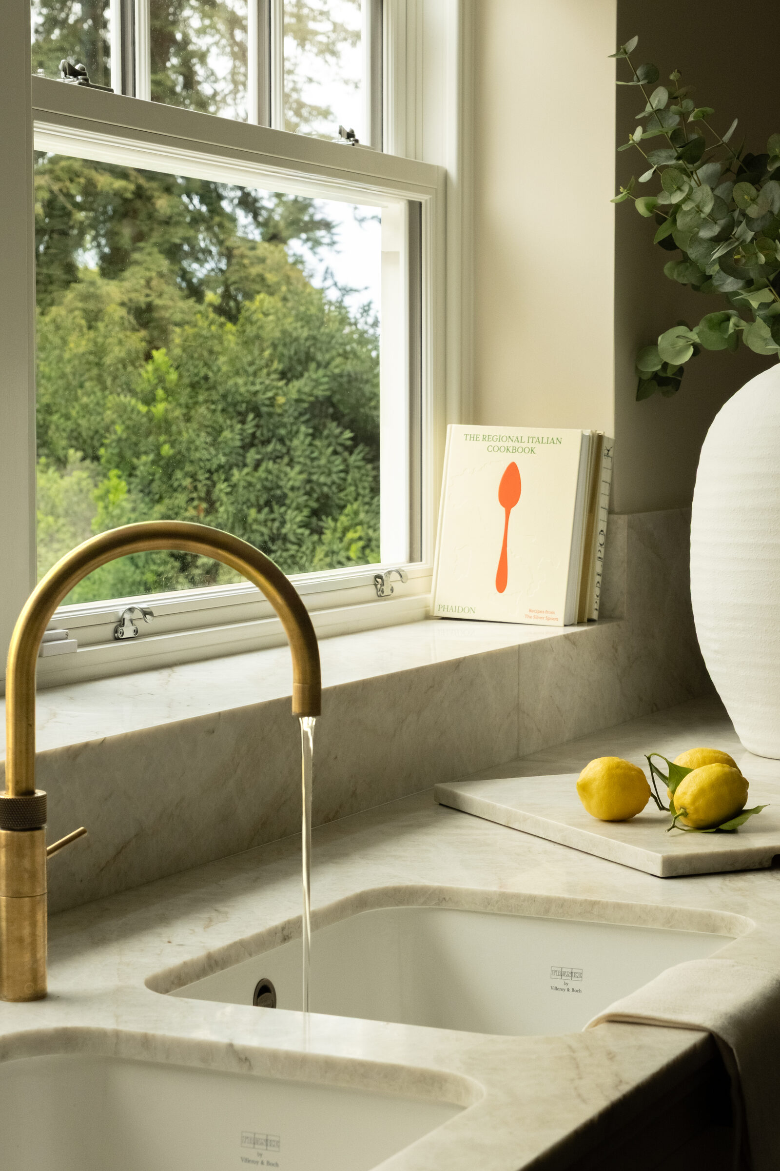 Unlacquered brass kitchen tap over a Taj Mahal quartzite sink with lemons and a cookbook on the sill